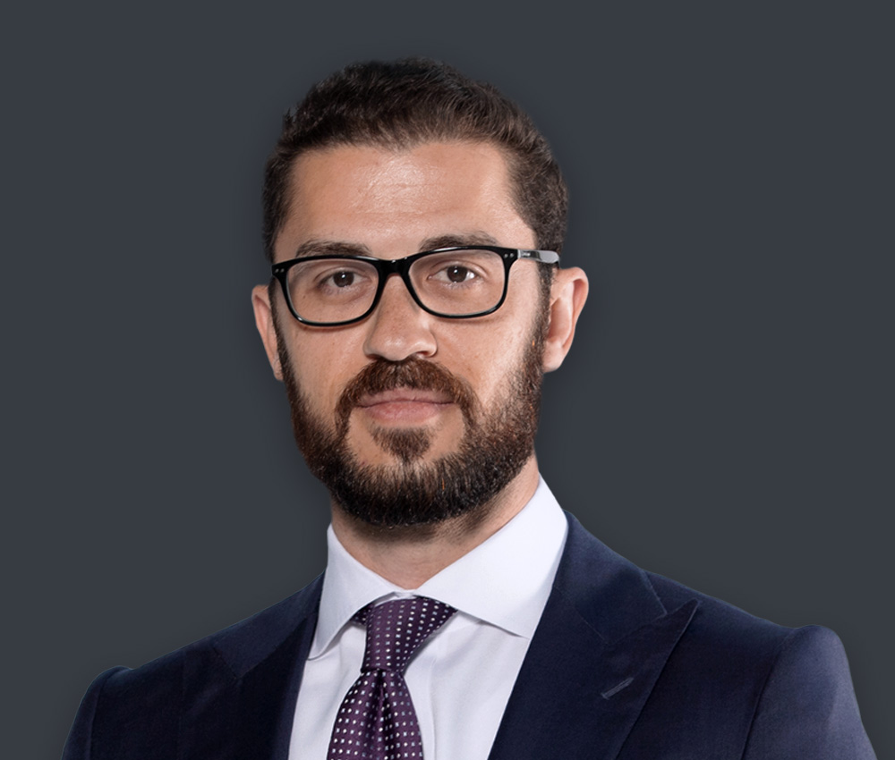 Portrait of a man with dark hair and glasses, wearing a navy suit and purple polka-dotted tie against a dark background.