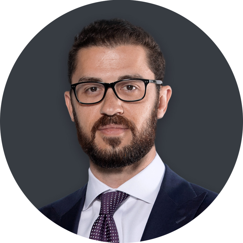 Professional headshot: man with dark hair and beard, glasses, navy suit, white shirt, and purple tie against a dark background.